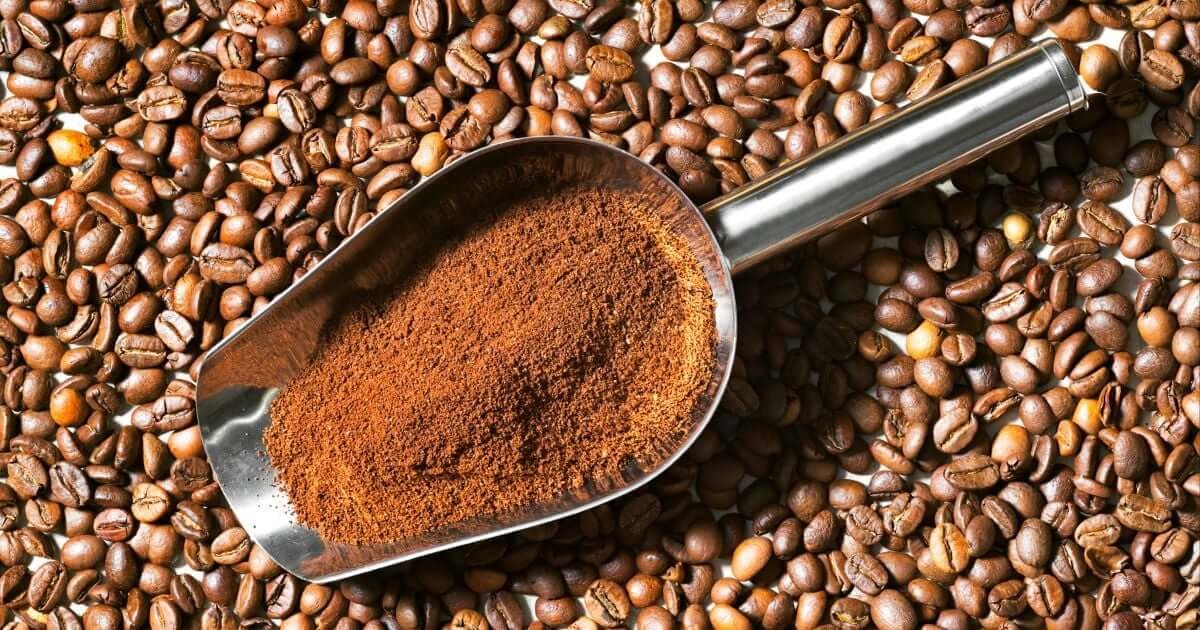 Hạt cà phê và bột cà phê trong muỗng kim loại, nền là nhiều hạt cà phê.
