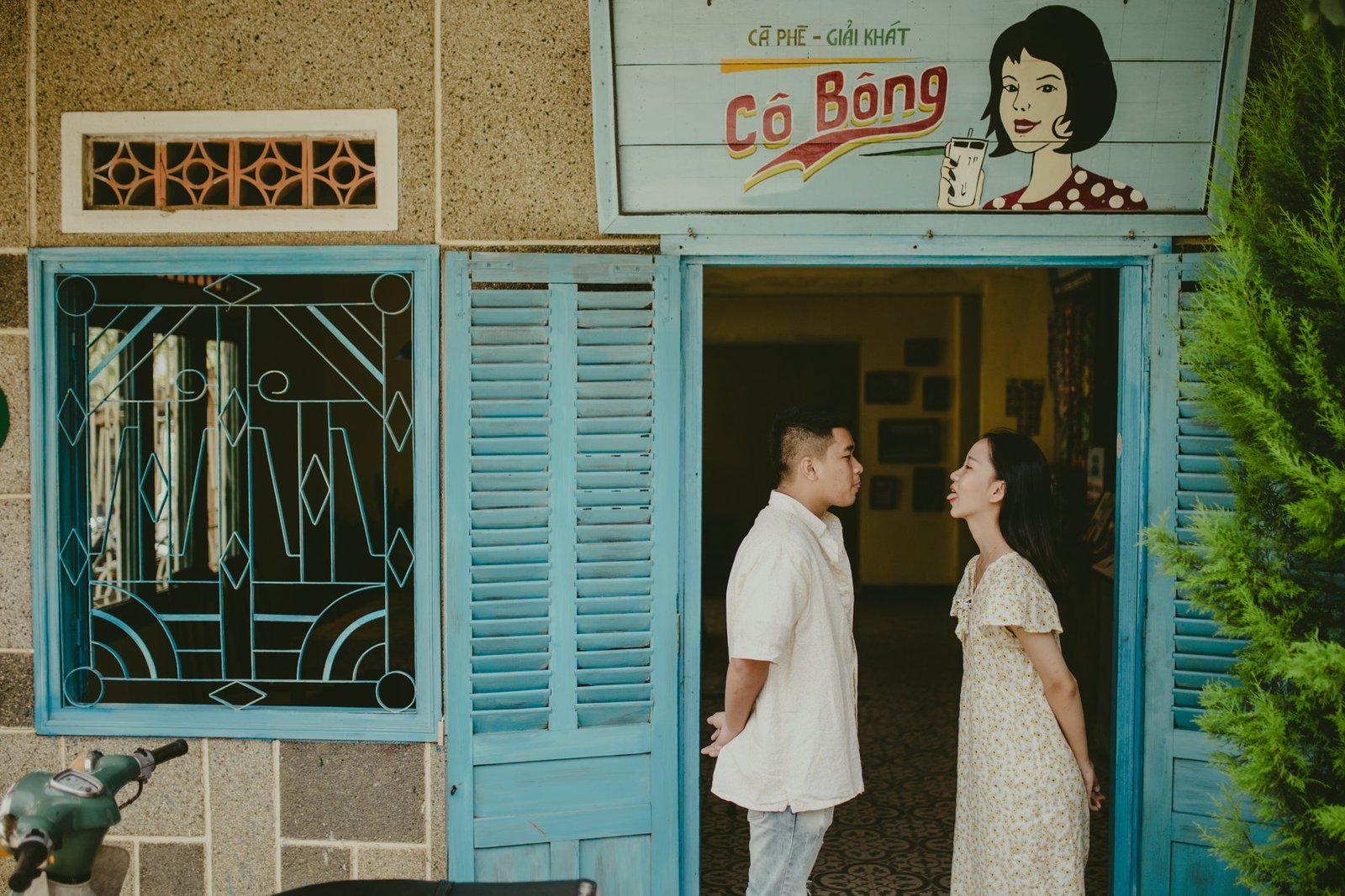 Hai người đứng đối diện nhau trước quán cà phê, cửa sổ và cửa ra vào màu xanh.