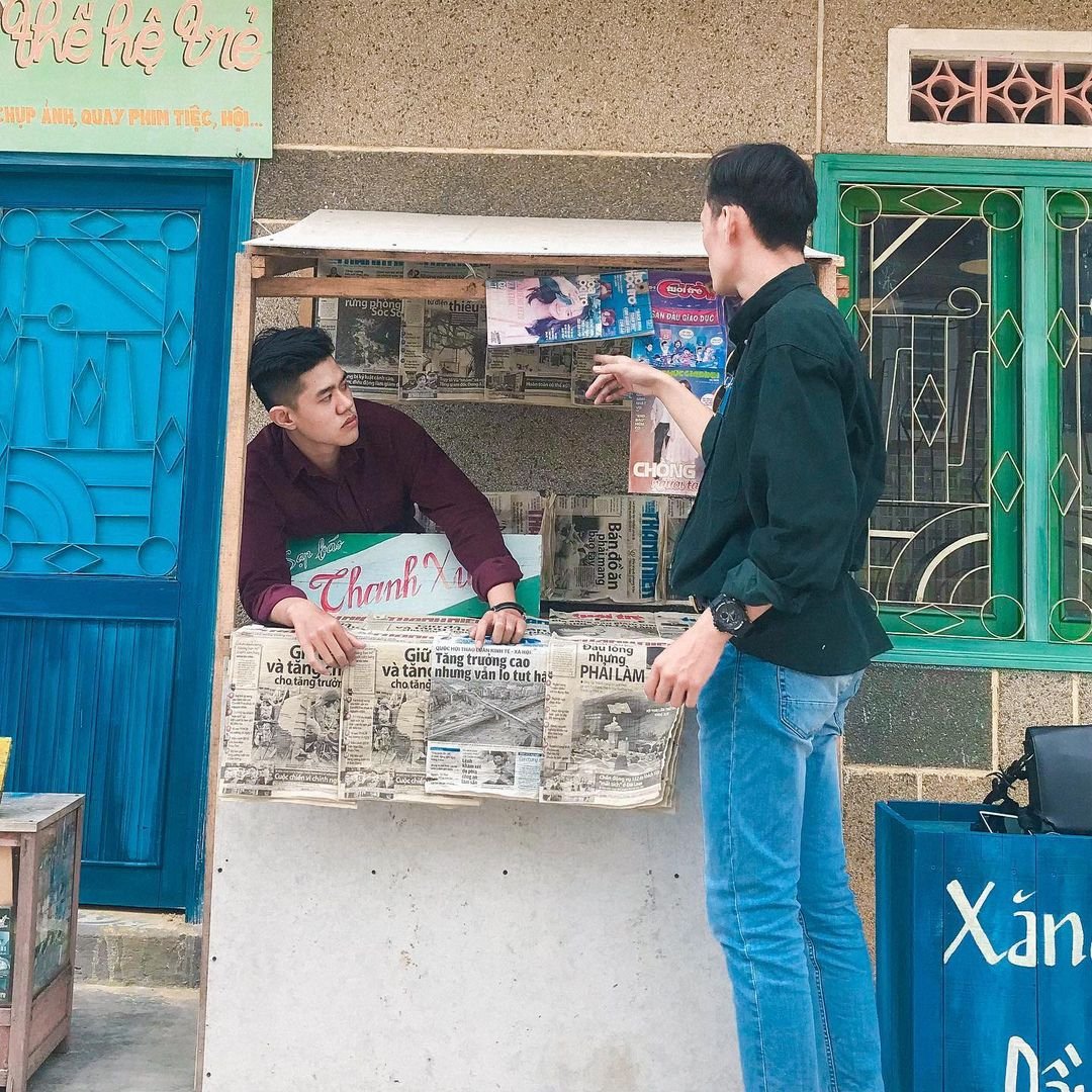Người đứng nói chuyện với người ngồi trong quầy báo nhỏ, xung quanh có các tờ báo và biển hiệu màu sắc.
