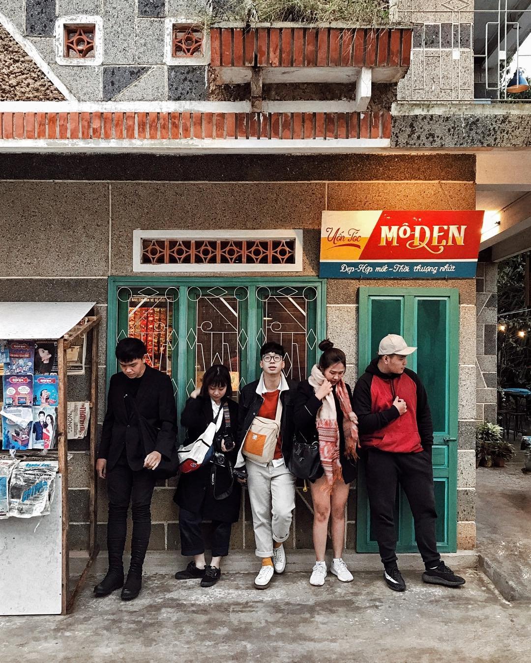 Nhóm người đứng trước cửa hàng với biển hiệu màu sắc rực rỡ, bên cạnh là sạp báo nhỏ.
