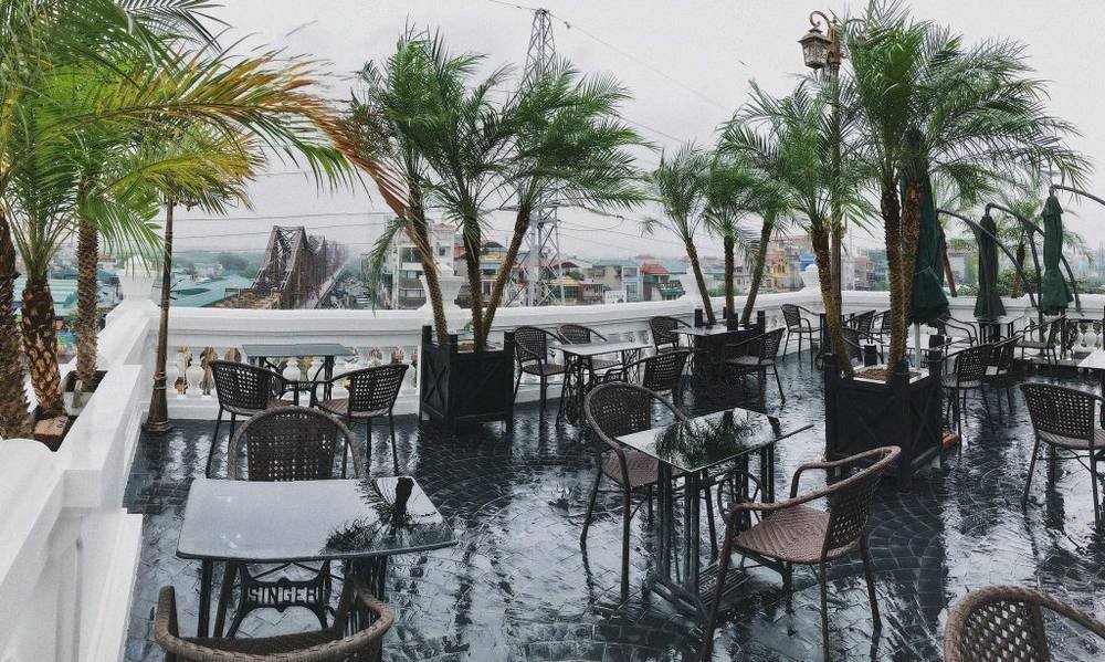Quán cà phê ngoài trời với bàn ghế và cây cọ, trời mưa, nhìn ra cảnh thành phố và cây cầu xa xa.
