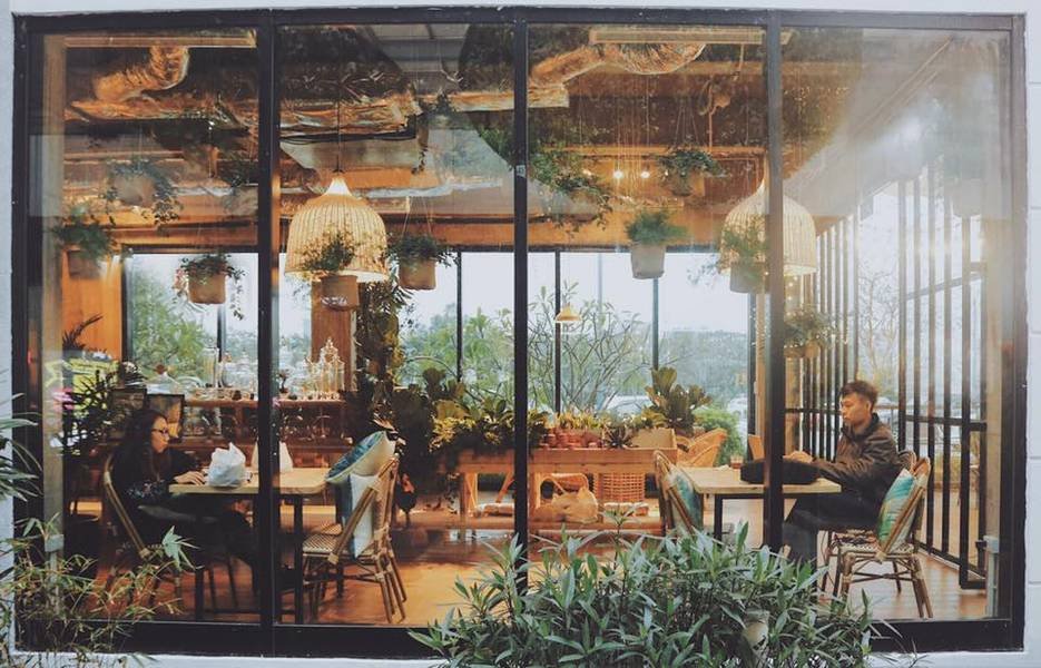 Quán cà phê với cửa kính lớn, bên trong có cây xanh và hai người đang ngồi thư giãn.